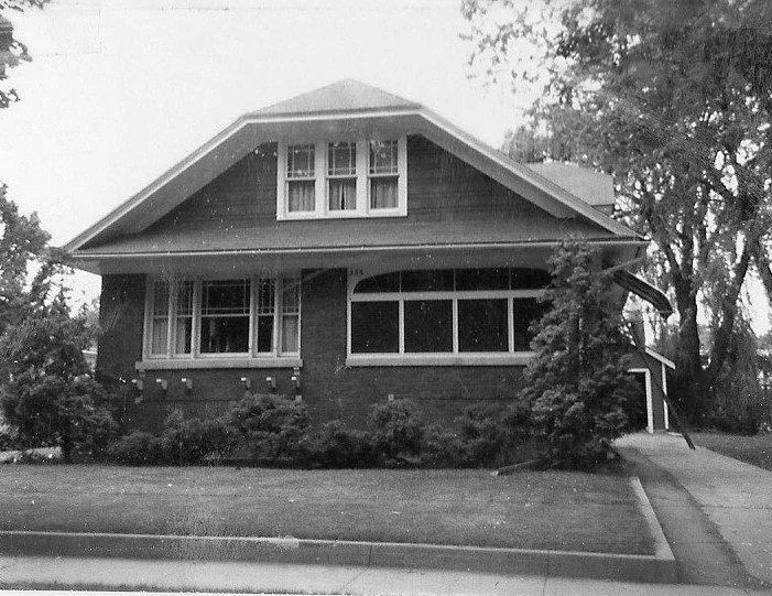 Black-and-white photo of a one-and-a-half-story brick bungalow with an enclosed front porch, dormer windows, driveway at right, and trees.