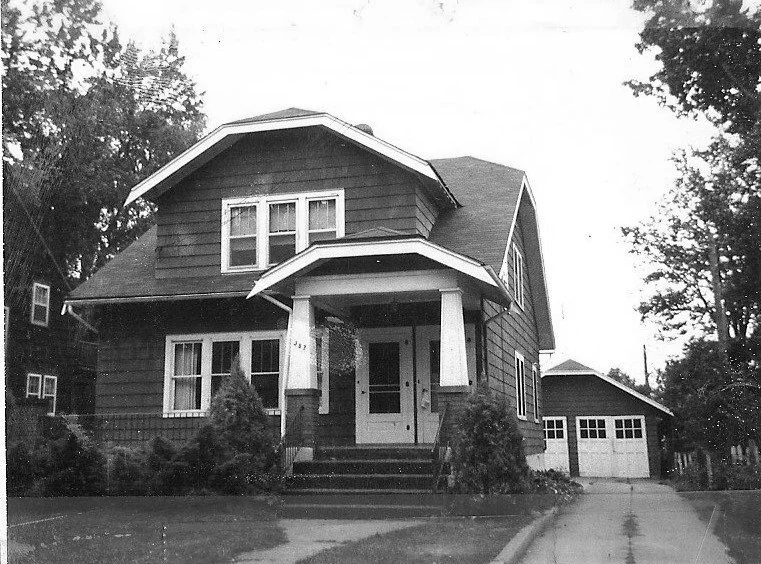Black-and-white photo of a two-story Craftsman-style house with a covered front porch supported by square columns, a driveway leading to a detached two-car garage, and trees framing the yard.