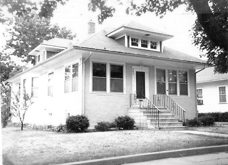 Black-and-white photo of a single-story brick bungalow with a front stairway and enclosed porch windows, dormer roof, and lawn with trees.