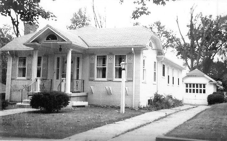 Black-and-white photo of a single-story bungalow with an enclosed front porch, central steps, and trimmed shrubs.