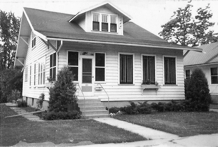 Black-and-white photo of a single-story bungalow with white siding, front steps and screened porch, a dormer window above, and shrubs lining the walkway.