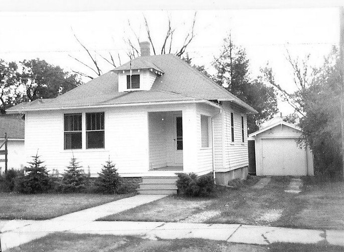 Black-and-white photo of a small single-story bungalow with a front porch, detached garage to the right, and a lawn with sidewalk in front.