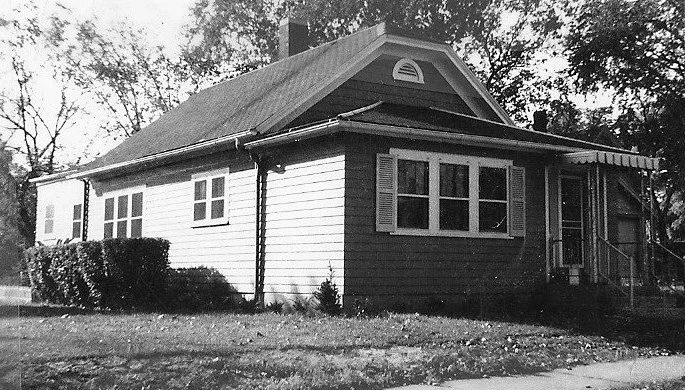 Black-and-white photo of a single-story bungalow with a small covered front porch, shuttered windows, and trees in the background.