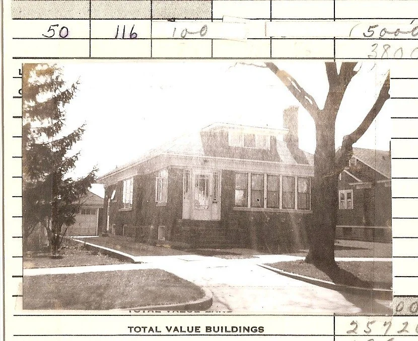 Black-and-white photo of a single-story brick bungalow with a front porch, large tree in the yard, driveway and detached garage; caption below reads "TOTAL VALUE BUILDINGS."