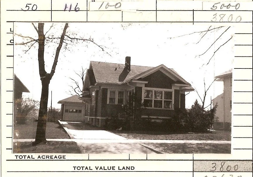 Black-and-white photo of a single-story bungalow with a front porch and driveway to a detached garage, flanked by leafless trees, mounted on a property assessment form with handwritten numbers.