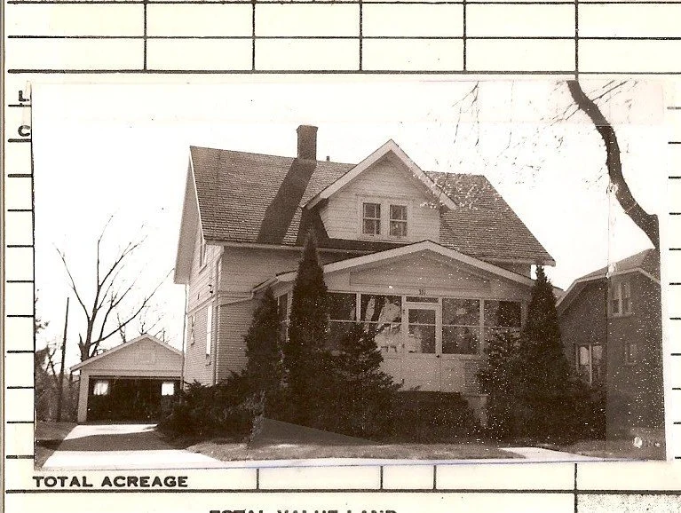 Black-and-white photo of a two-story gabled house with enclosed front porch, tall shrubs, driveway leading to a detached garage at left; archival grid border reading "TOTAL ACREAGE."