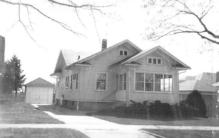 Black-and-white photo of a single-story Craftsman-style house with an enclosed front porch, driveway leading to a detached garage, and bare tree branches framing the scene.