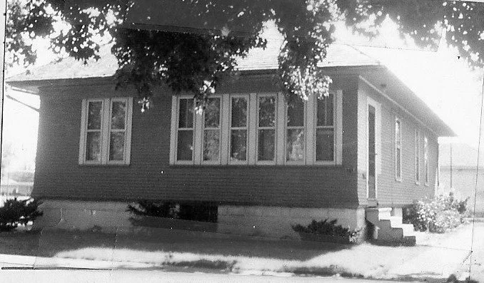 Black-and-white photo of a single-story wood-frame bungalow with a row of tall windows, small front steps, and tree branches overhead.
