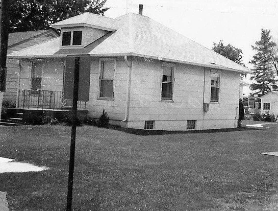 Black-and-white photo of a single-story detached house with a low-pitched roof and small dormer, front porch, and a grassy lawn with a signpost in the foreground.
