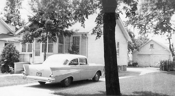 Black-and-white photo of a vintage sedan parked in a driveway beside a small house, with a tree in the foreground and a detached garage in the background.