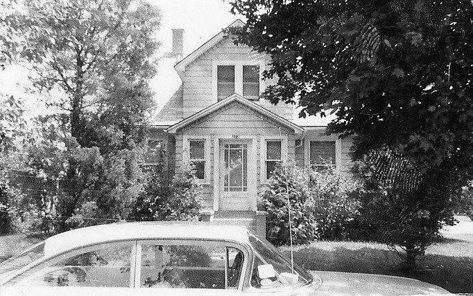 Black-and-white photo of a two-story house with a small front porch, trees and shrubs around it, and a vintage car parked in front.