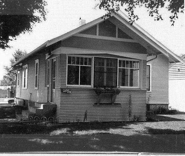 Black-and-white photo of a single-story Craftsman-style bungalow with an enclosed front porch, large multi-pane windows, wood siding, a small window box, and trees casting shadows.