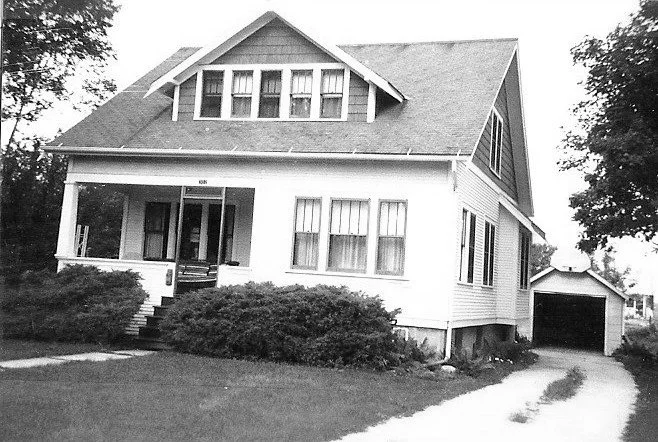 Black-and-white photo of a two-story Craftsman-style house with a covered front porch, upper dormer windows, shrubs in front, and a driveway leading to a detached garage.