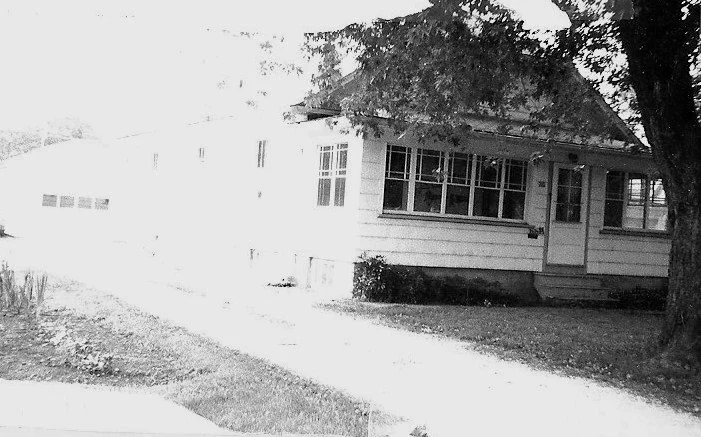 Black-and-white photo of a single-story house with an enclosed front porch, large tree at right, and a driveway leading to a detached garage.
