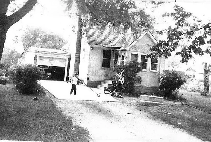 Black-and-white photo of a small bungalow with detached garage; a child on the driveway holding a long stick beside a fallen bicycle near the front steps, trees and bushes frame the yard.