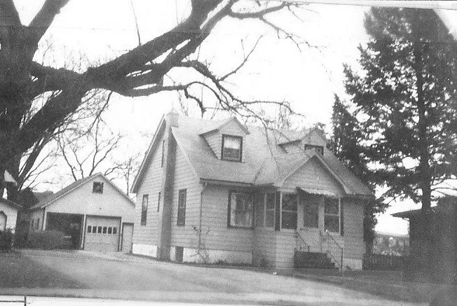Black-and-white photo of a two-story house with dormer windows and a front porch, a detached garage to the left, and large bare tree branches framing the scene.
