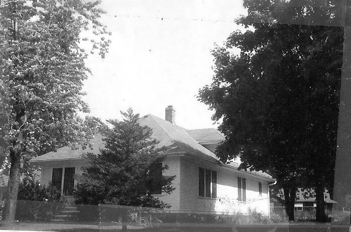 Black-and-white photo of a single-story house with a hipped roof and chimney, surrounded by large trees and a small front porch.