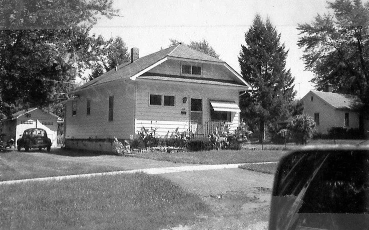 Black-and-white photo of a single-story bungalow with a small front porch and awning, lawn and driveway with a parked car and garage.
