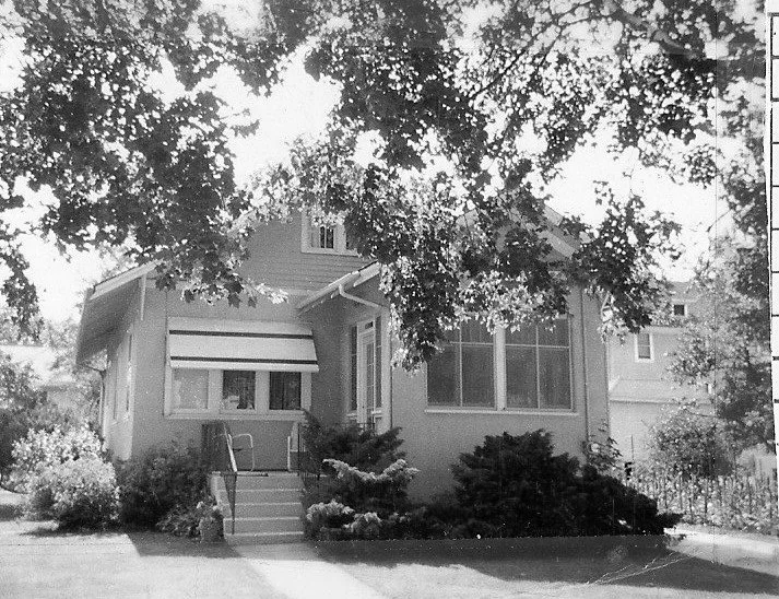 Black-and-white photo of a single-story bungalow with front steps, window awning, shrubs, and large tree branches overhead.