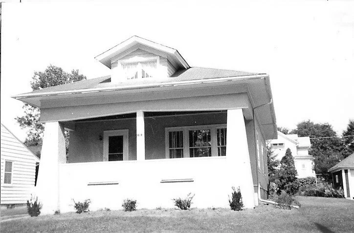 Black-and-white photo of a single-story bungalow with a covered front porch, square columns, dormer window, and small front lawn.