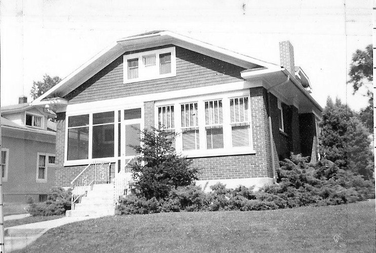 Black-and-white photo of a one-story brick bungalow with a screened front porch, front steps, window blinds, and shrubs on a grassy lawn.