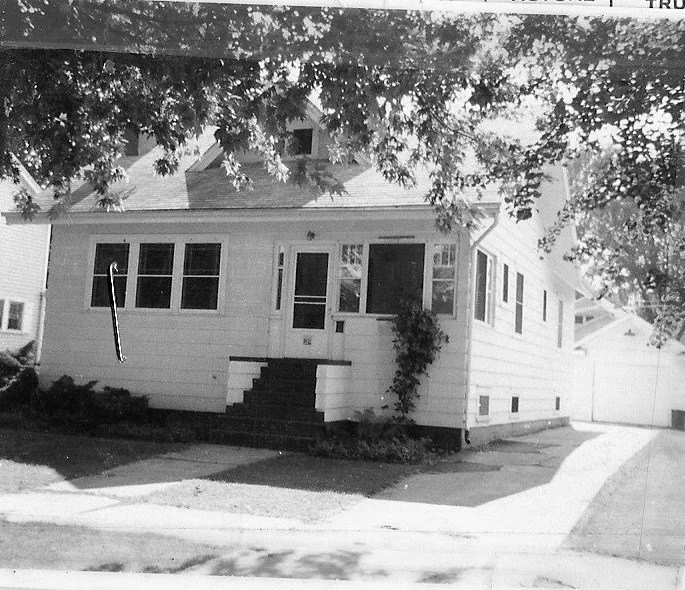 Black-and-white photo of a small single-family house with a screened front porch, center steps leading up to the door, a driveway on the right, and tree branches overhanging the yard.