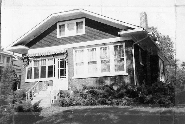 Black-and-white photo of a single-story brick bungalow with an enclosed front porch, awning, front steps, and shrubs.