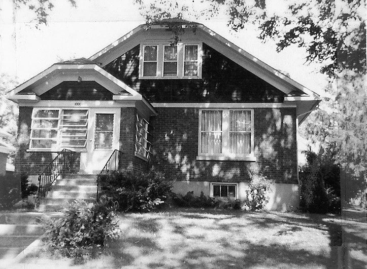 Black-and-white photo of a brick bungalow with a screened front porch, front steps and a dormer window, shaded by trees.