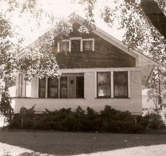 Vintage black-and-white photo of a small bungalow with an enclosed front porch, shrubs, and overhanging trees.