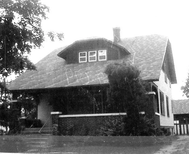 Black-and-white photo of a Craftsman-style bungalow with a wide covered front porch, three small dormer windows, and overgrown shrubs partially obscuring the entrance.