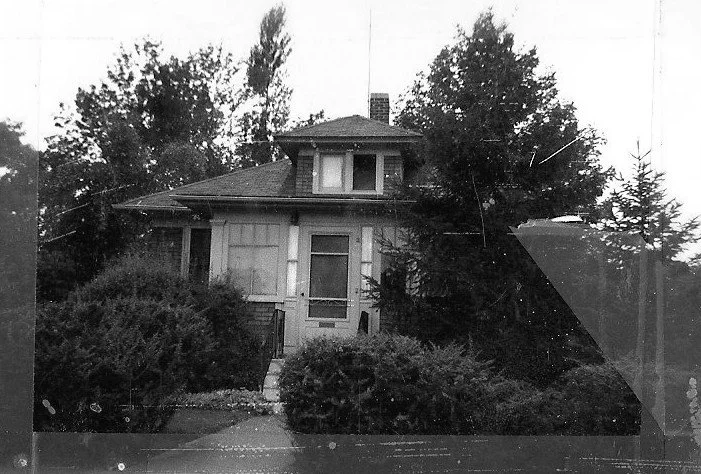 Black-and-white photo of an older two-story house partially obscured by overgrown bushes and trees.