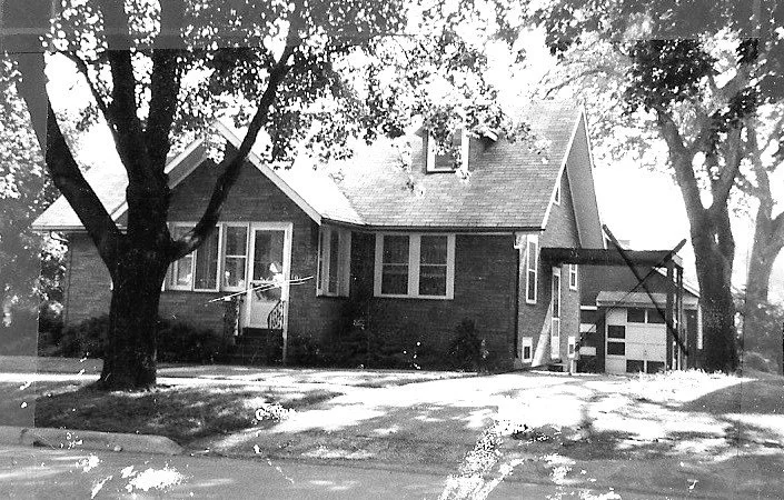 Black-and-white photo of a single-story brick house with front porch, large trees, driveway, and detached garage.
