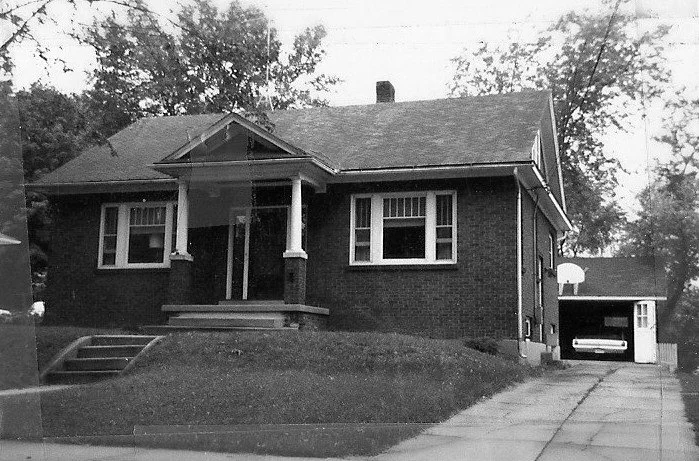 Black-and-white photo of a one-story brick bungalow with a small front porch and steps, lawn, driveway leading to a detached garage with a car inside.