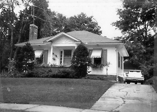 Black-and-white photo of a small single-story suburban house with a front porch, window awnings, tall trees behind, and a vintage car parked in the driveway.