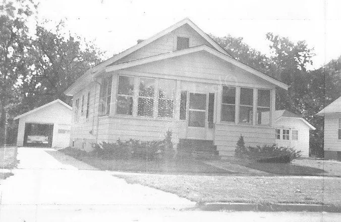 Black-and-white photo of a single-story bungalow with an enclosed front porch, concrete driveway leading to a detached garage at left, and low shrubs in front.