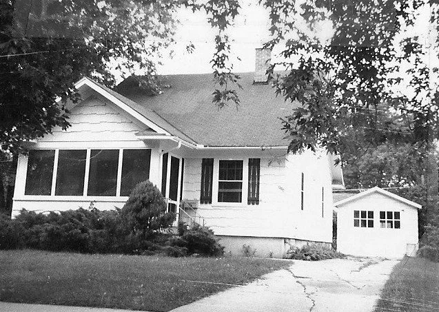 Black-and-white photo of a one-story bungalow with an enclosed front porch, driveway leading to a detached garage, and trees framing the foreground.