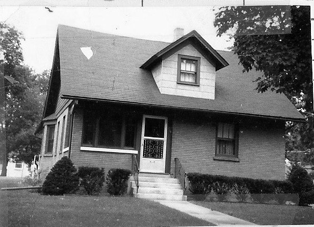 Black-and-white photo of a one-and-a-half-story brick bungalow with a front porch, central entry with steps and decorative screen door, dormer window, and shrubs lining the lawn.