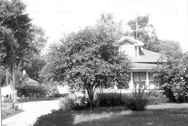 Black-and-white photo of a bungalow-style house partly hidden by a large tree in the front yard, with lawn and driveway visible.