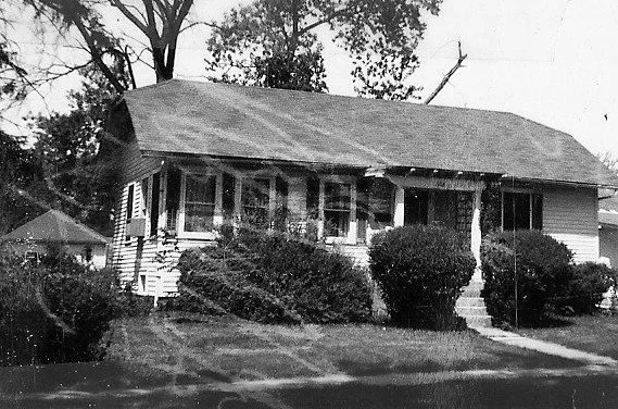 Black-and-white photo of a single-story bungalow with a covered front porch, concrete steps and large shrubs lining the front yard, set beneath tall trees.