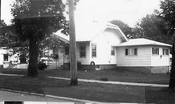 Black-and-white photo of a single-story house with an attached garage, front lawn, sidewalk and trees.