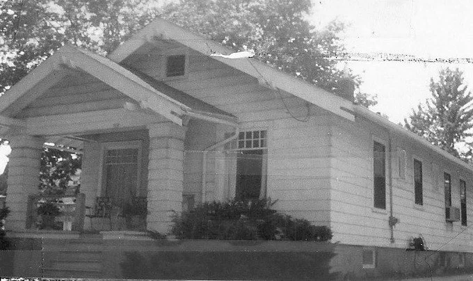 Black-and-white photo of a single-story bungalow with a covered front porch, square columns, shrubs, and trees in the background.
