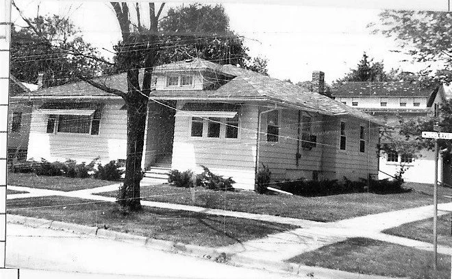 Black-and-white photo of a single-story bungalow on a corner lot with a tree in front, sidewalks and neighboring houses in the background.