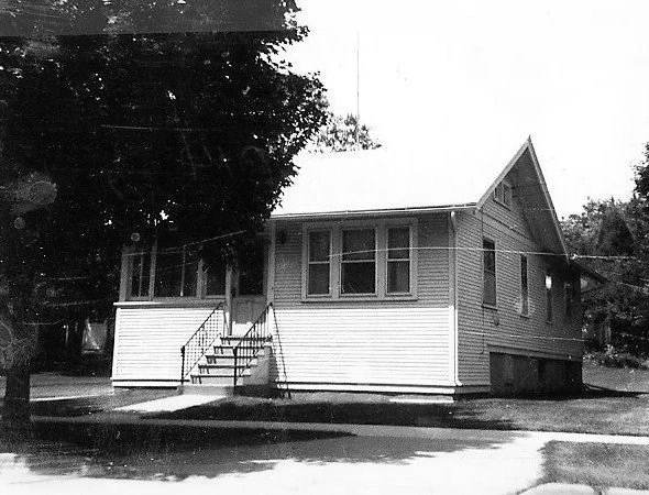 Black-and-white photo of a small one-story clapboard house with a front porch, central stairs, and a large tree at the left.