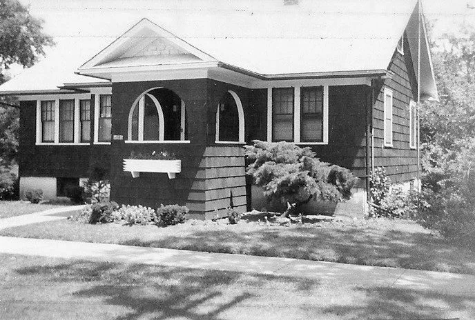 Black-and-white photo of a single-story bungalow with a covered front porch featuring arched openings, surrounding shrubs, and a sidewalk in the foreground.