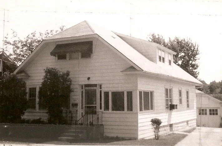 Black-and-white photo of a two-story wood-sided bungalow with a small enclosed front porch, dormer windows, shrubs by the entrance, and a detached garage at the right.