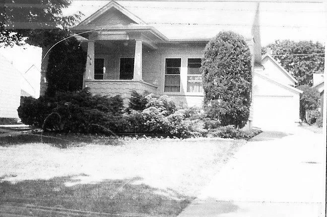 Black-and-white photo of a small bungalow with a covered front porch, dense shrubs and a lawn, and a driveway leading to a detached garage.