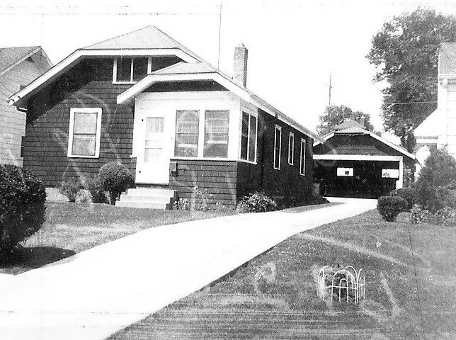 Black-and-white photo of a single-story bungalow with an enclosed front porch, long driveway leading to a detached garage, and a lawn with shrubs.