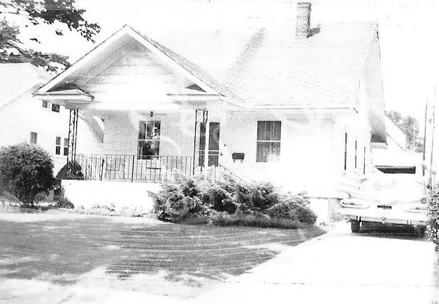 Small single-story white bungalow with a covered front porch, shrubs and lawn, and a driveway with a parked car on the right.