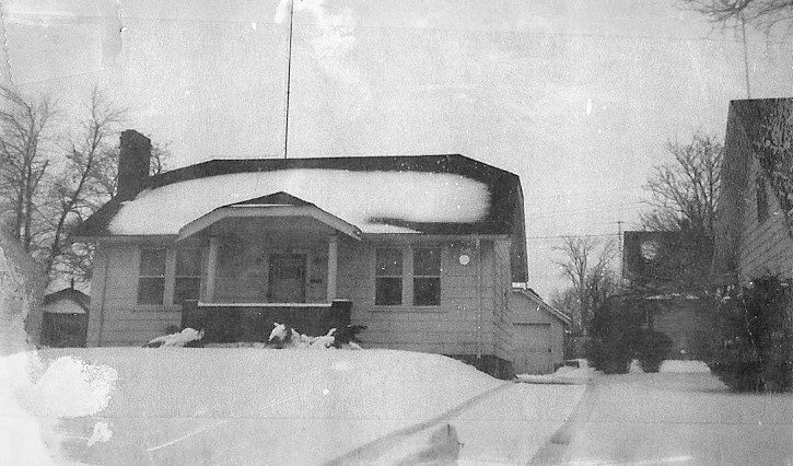Black-and-white photo of a one-story bungalow with a snow-covered roof and yard, a small front porch, and a cleared driveway leading to a detached garage.