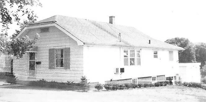 Small single-story white house with horizontal siding, shuttered front window, window air-conditioner, chimney and trees in the background.